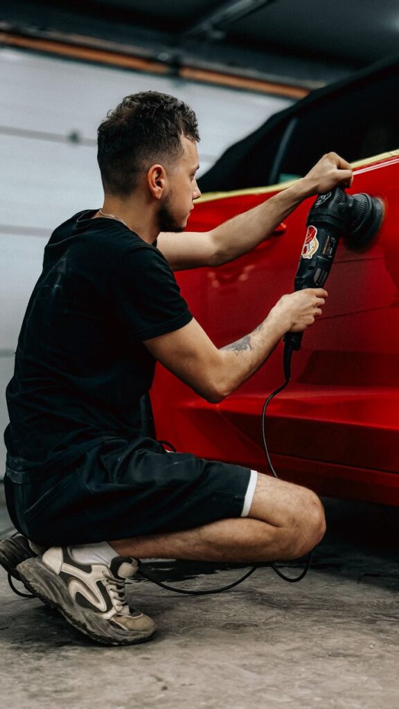 A man in casual attire meticulously polishing a red car in a garage setting, focusing on auto detailing.